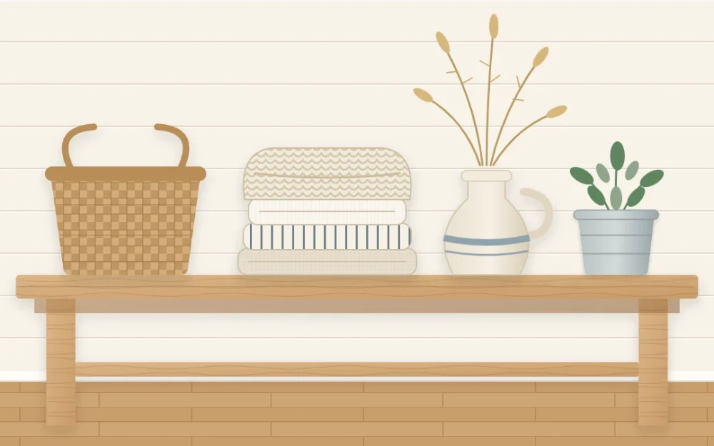 Wooden bench against a pale plank wall holding folded linen and a chunky knitted throw, a woven basket, a stoneware jug of dried stems and a small galvanized pot with a plant
