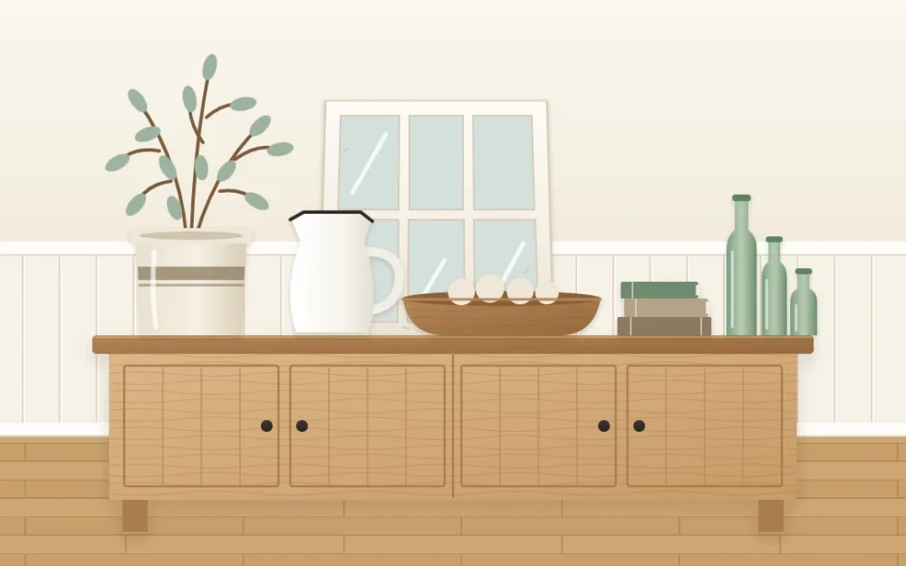 Wooden sideboard styled with a stoneware crock of branches, a white enamel-style pitcher, a long wooden bowl, three green glass bottles, old books and a leaning window-style frame